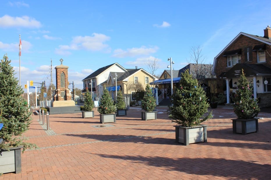 A public square with potted trees, benches, and surrounding buildings under a clear sky.