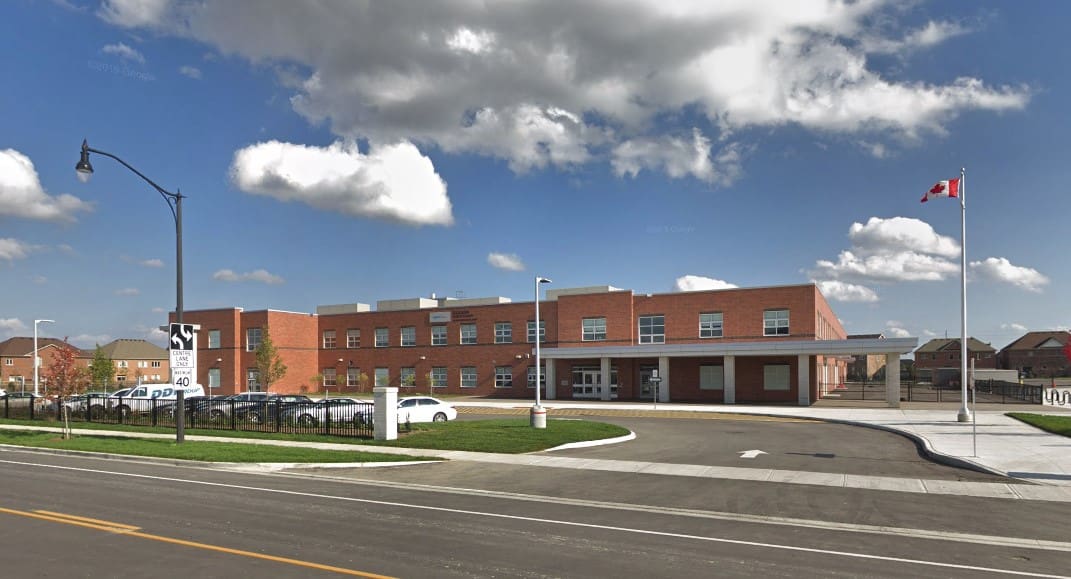 A modern redbrick school building under a cloudy sky, with a Canadian flag flying, surrounded by a residential area and parked cars.