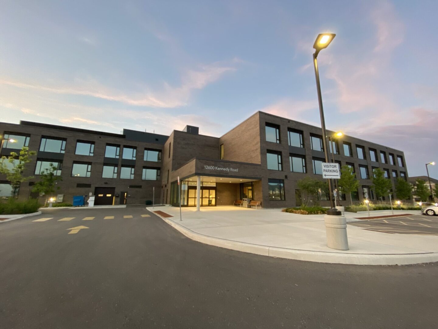 A modern three-story school building at dusk, with an illuminated parking area and a sign indicating "Visitor Entrance" at the front.