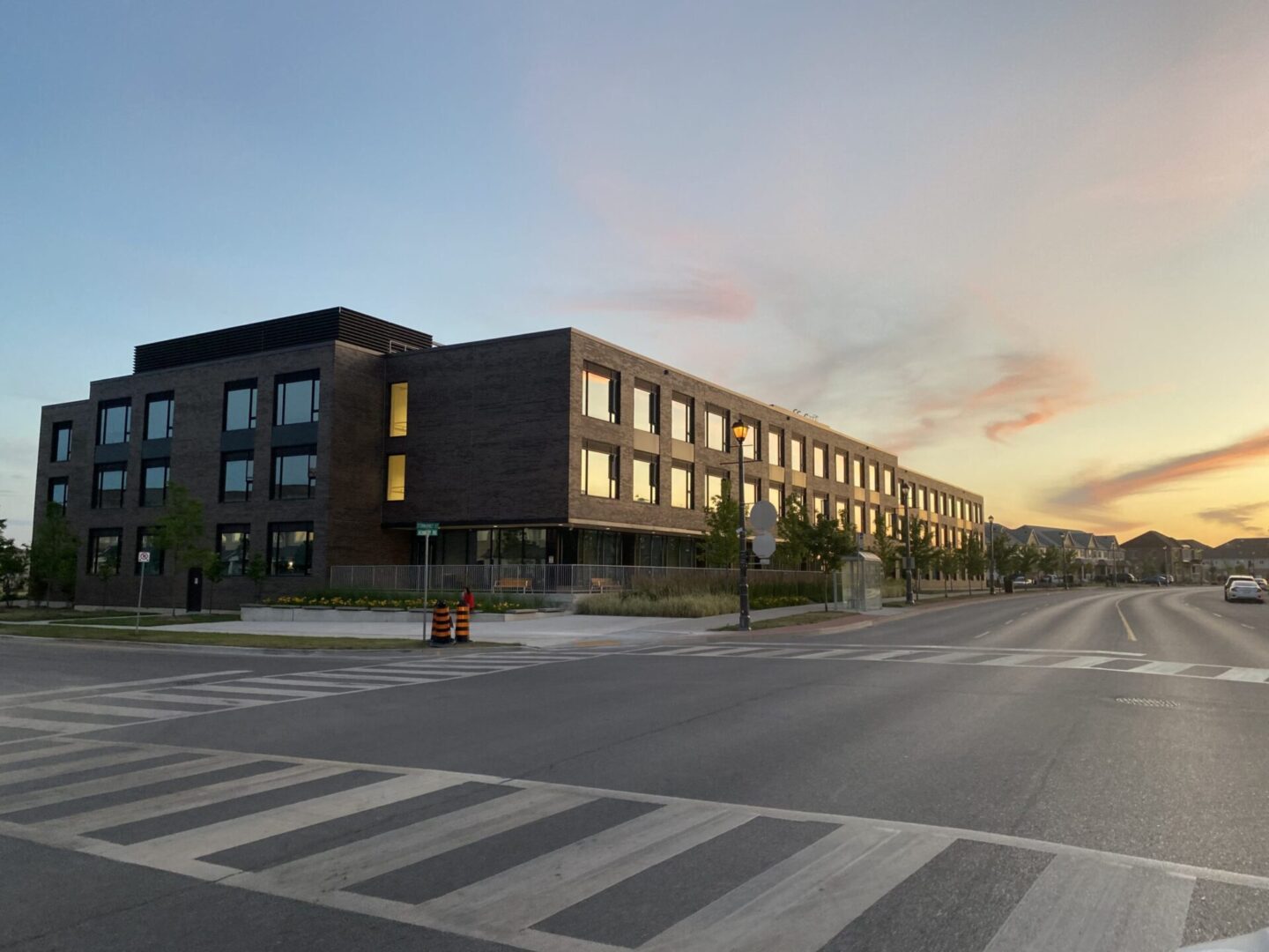 Modern two-story building at dusk with illuminated windows, situated beside a street with a crosswalk under a colorful sunset sky.