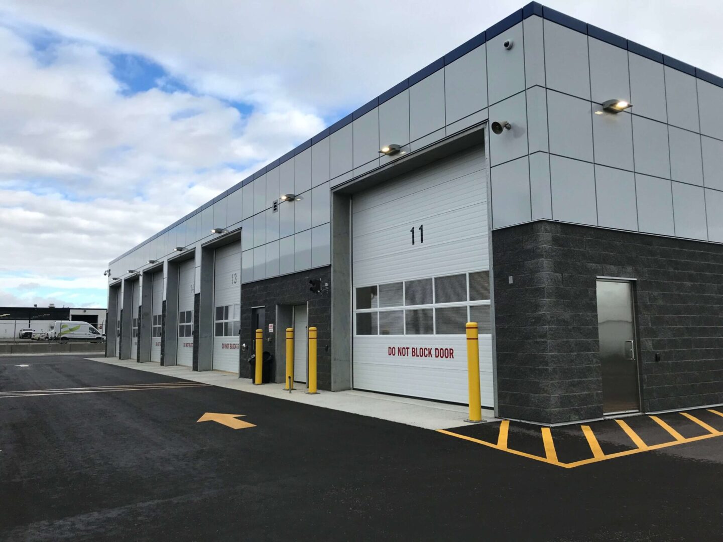 Modern industrial warehouse exterior with multiple numbered loading bays, marked pavement, and cloud-filled sky.