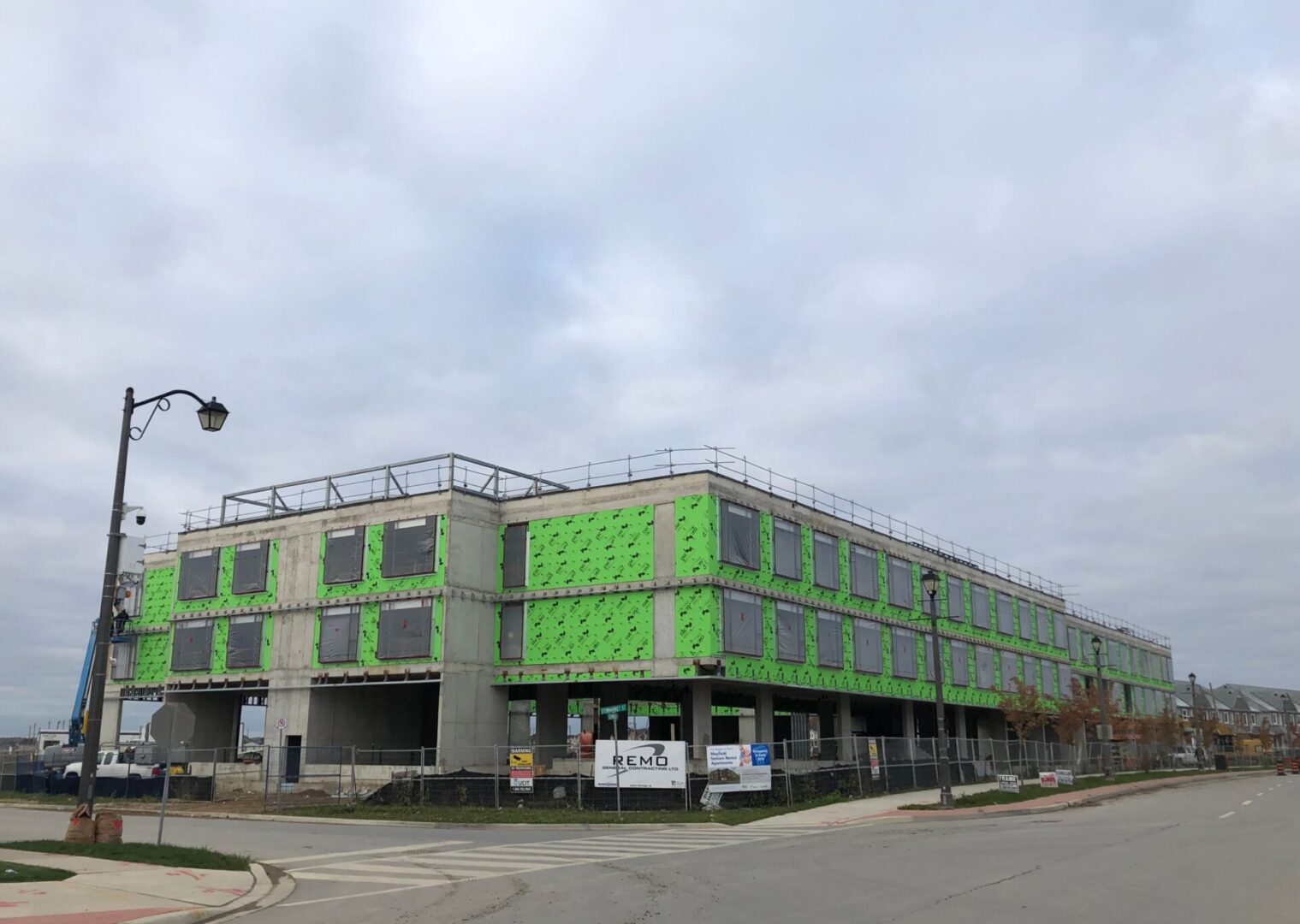 A modern building under construction covered with green weather-resistant sheathing, surrounded by a paved road and a cloudy sky.