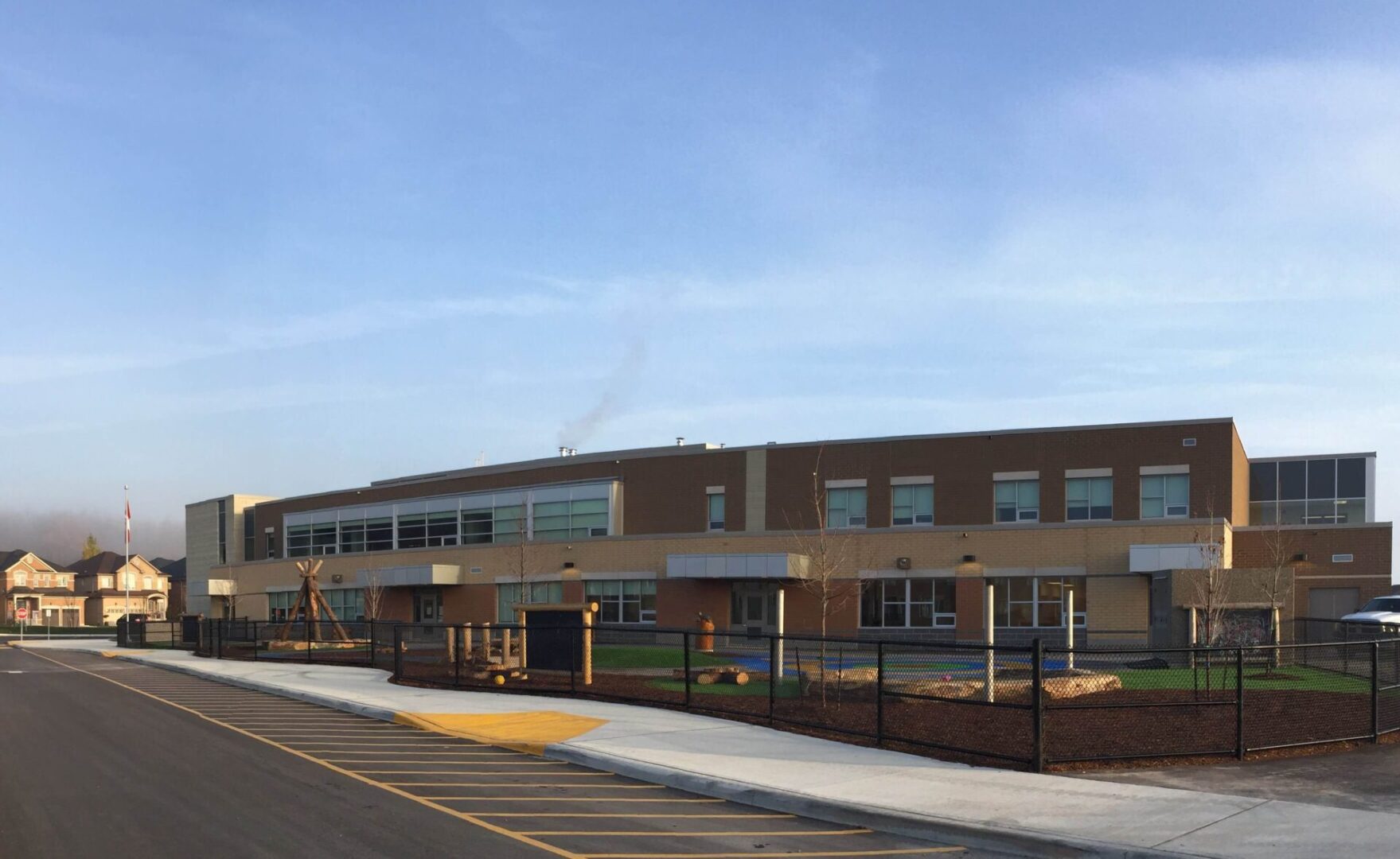 Modern two-story school building with a beige and brown facade, playground in the foreground, and a flagpole, under a clear sky at sunrise.