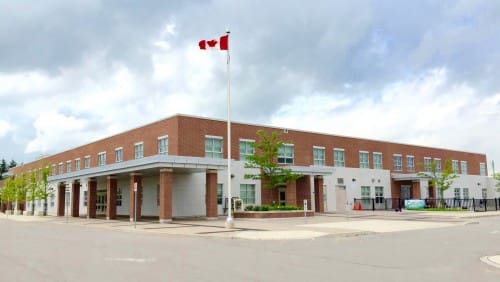 A two-story brick school building with a flagpole displaying the Canadian flag in front, under a partly cloudy sky.