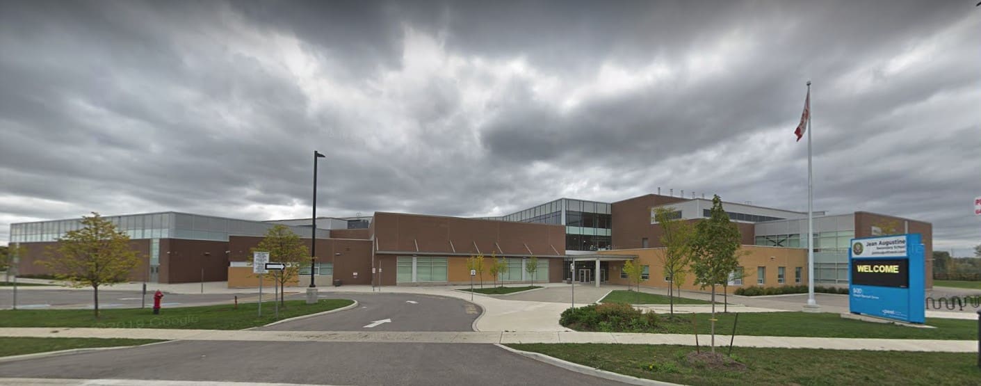 A modern school building under an overcast sky, featuring large windows, a curved driveway, and a flagpole with a Canadian flag.