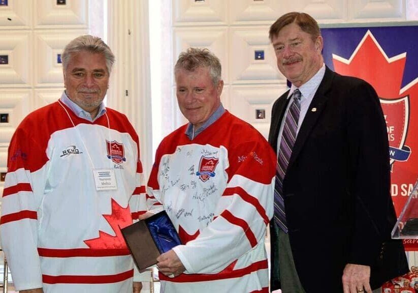 Three men standing together indoors, two in white and red Canada themed hockey jerseys and one in a suit, holding a sports memorabilia item.