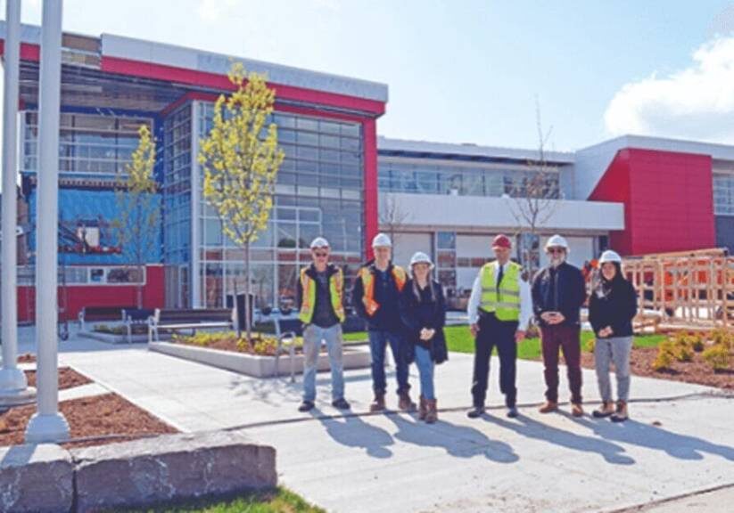 Group of six construction workers wearing hard hats and safety vests standing in front of a modern building with landscaping.